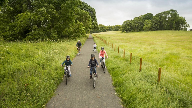 A famly cycling on a tarmac track on the Dragon cycle trail, at Wallington, Northumberland, with grassy fields on both sides, trees on the left and a fence on the right.